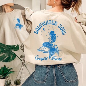 Peut inclure: Un sweat-shirt beige clair avec un graphique bleu d'une sir&egrave;ne portant un chapeau de cow-boy. Le texte "Saltwater Soul Cowgirl Heart" est imprim&eacute; sur le sweat-shirt.