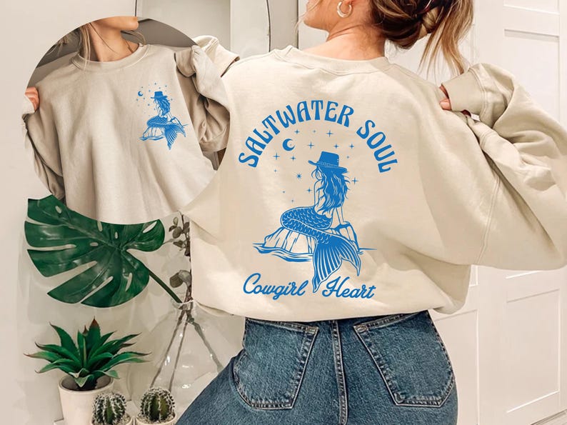 Peut inclure: Un sweat-shirt beige clair avec un graphique bleu d'une sir&egrave;ne portant un chapeau de cow-boy. Le texte "Saltwater Soul Cowgirl Heart" est imprim&eacute; sur le sweat-shirt.