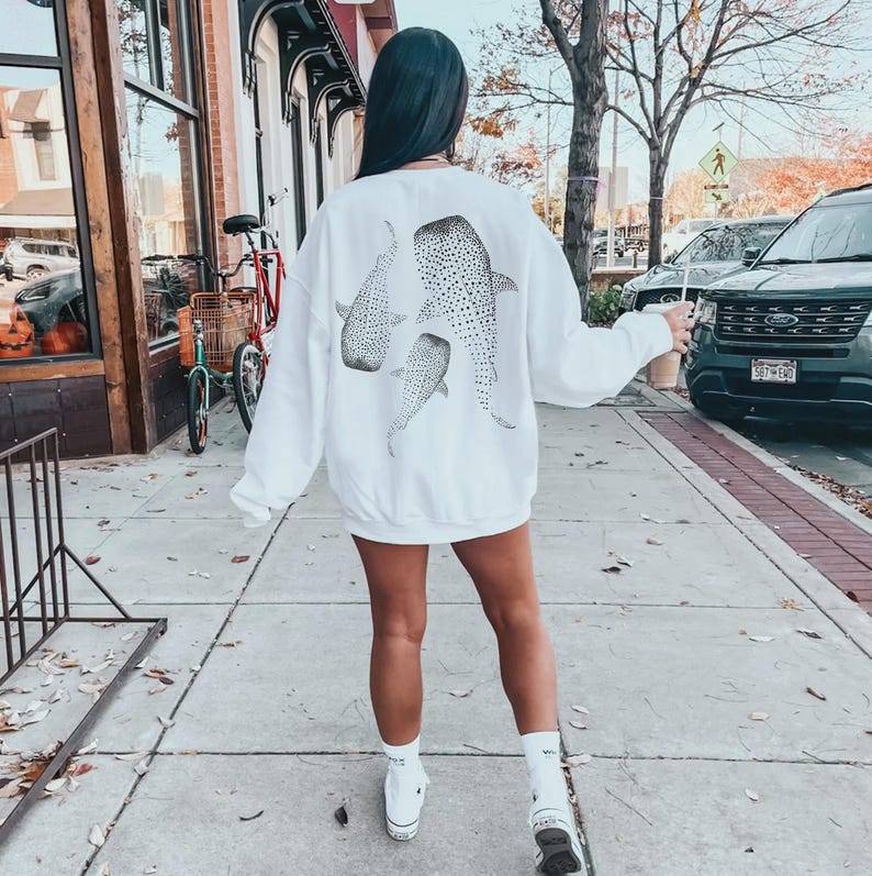 May include: White sweatshirt featuring three whale shark illustrations in a dotted pattern. The sweatshirt is a relaxed fit, perfect for casual wear. The design is centered on the back of the garment.