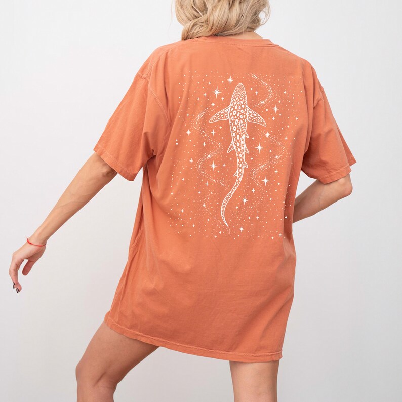 Puede incluir: Camiseta naranja quemado con una ilustraci&oacute;n blanca de un tibur&oacute;n con un patr&oacute;n de puntos y una cola larga, rodeado de estrellas. La camiseta de manga corta tiene un corte relajado, ideal para el uso diario.