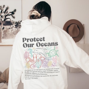 Könnte beinhalten: Weißer Hoodie mit einem Grafikdruck eines Delfins, einer Schildkröte, eines Kraken und eines Korallenriffs. Der Text "Protect Our Oceans" ist in Grau auf dem Hoodie gedruckt.