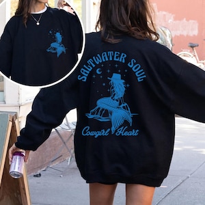 Peut inclure: Sweat-shirt noir avec un graphique bleu d'une sir&egrave;ne avec le texte "Saltwater Soul Cowgirl Heart" au dos.