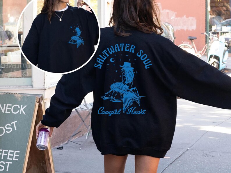 Peut inclure: Sweat-shirt noir avec un graphique bleu d'une sir&egrave;ne avec le texte "Saltwater Soul Cowgirl Heart" au dos.