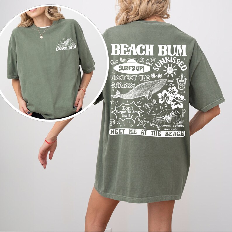 May include: An olive green t-shirt featuring a white graphic on the front and back. The back graphic includes the words "BEACH BUM" and "SUNKISSED", alongside illustrations of a whale, sunglasses, and other beach-themed elements. The shirt has a loose fit.