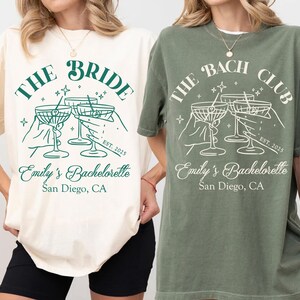 May include: Two white t-shirts with green text and graphics. The text on the left shirt reads "The Bride" and the text on the right shirt reads "The Bach Club". Both shirts feature a graphic of four hands holding up martini glasses. The text "Emily's Bachelorette San Diego, CA" appears below the graphic on both shirts.