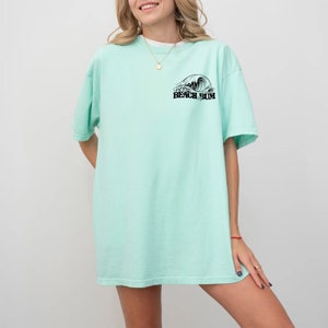 May include: A pale green t-shirt featuring a black wave graphic and the text "Beach Bum". The shirt has a loose fit and short sleeves. The model is wearing a gold necklace.