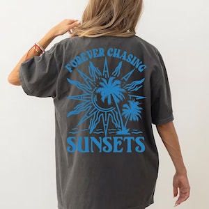 Puede incluir: Una camiseta gris oscuro con un estampado gr&aacute;fico azul de un sol, palmeras y el texto "FOREVER CHASING SUNSETS".