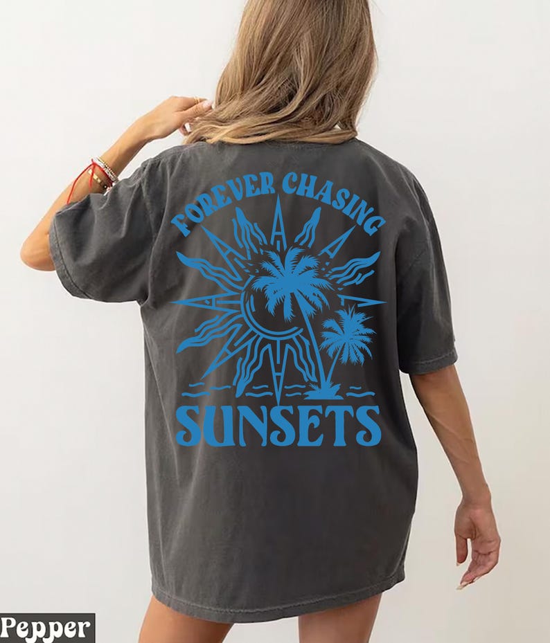 Puede incluir: Una camiseta gris oscuro con un estampado gr&aacute;fico azul de un sol, palmeras y el texto "FOREVER CHASING SUNSETS".