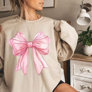 May include: A beige sweatshirt with a large pink bow graphic printed on the front.