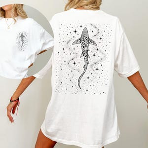 Puede incluir: Camiseta blanca con una ilustraci&oacute;n en blanco y negro de un tibur&oacute;n con un patr&oacute;n de puntos, rodeado de estrellas y un rastro punteado. El dise&ntilde;o est&aacute; en la espalda y una versi&oacute;n m&aacute;s peque&ntilde;a en el pecho izquierdo.