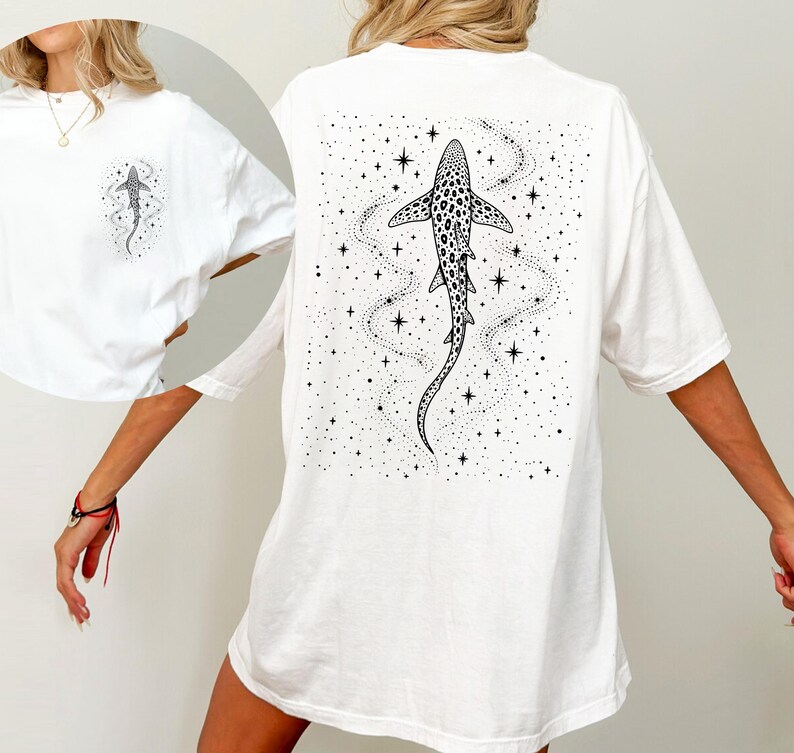 Puede incluir: Camiseta blanca con una ilustraci&oacute;n en blanco y negro de un tibur&oacute;n con un patr&oacute;n de puntos, rodeado de estrellas y un rastro punteado. El dise&ntilde;o est&aacute; en la espalda y una versi&oacute;n m&aacute;s peque&ntilde;a en el pecho izquierdo.