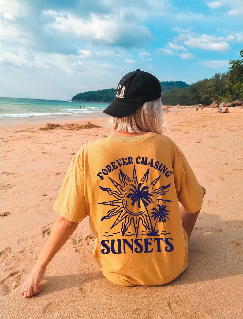 Puede incluir: Una camiseta amarilla con un gr&aacute;fico de un sol, palmeras y el texto "FOREVER CHASING SUNSETS".