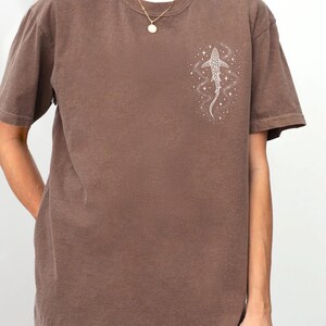 Puede incluir: Camiseta color espresso con un gr&aacute;fico blanco de un tibur&oacute;n ballena y un dise&ntilde;o estrellado. La camiseta de manga corta tiene cuello redondo y un corte relajado. La modelo lleva una camiseta interior blanca y un collar dorado.