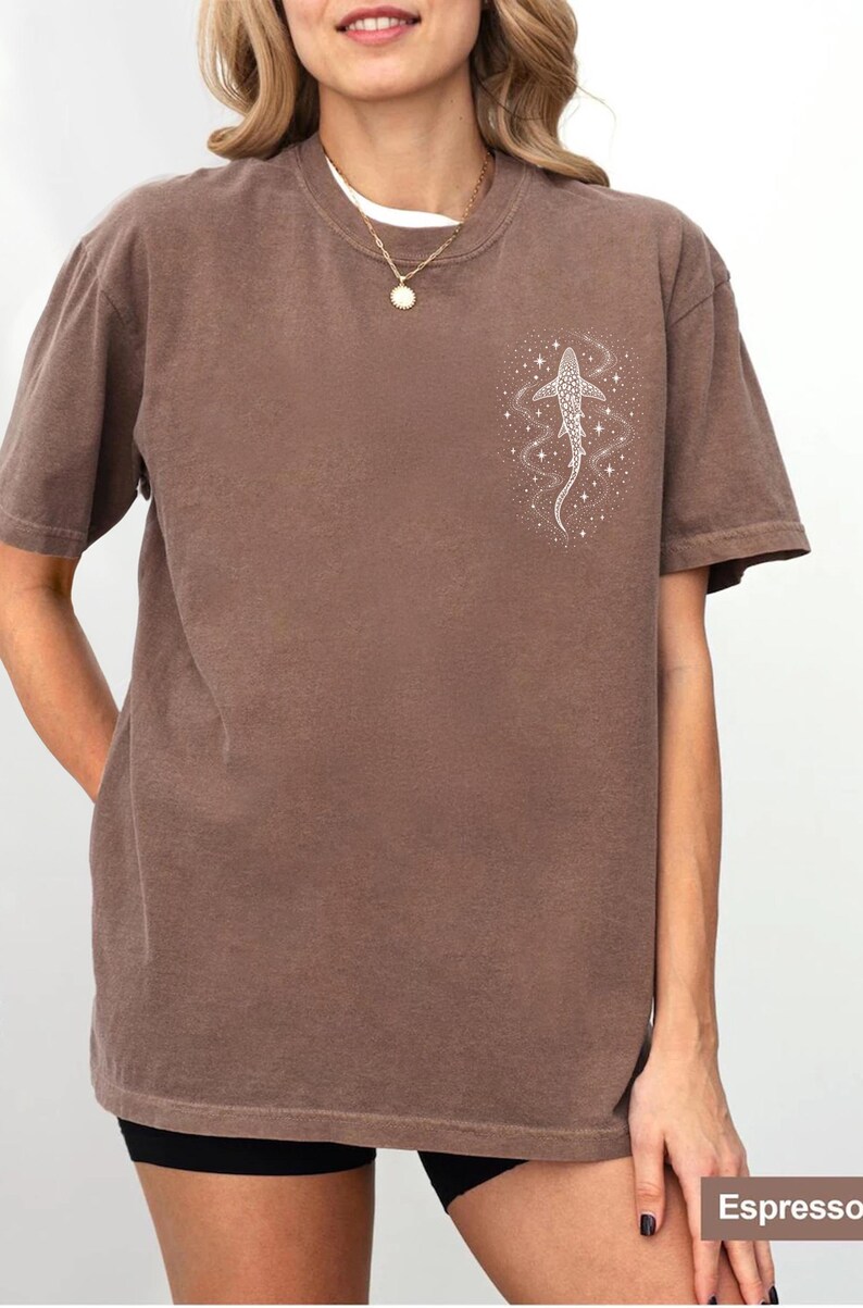 Puede incluir: Camiseta color espresso con un gr&aacute;fico blanco de un tibur&oacute;n ballena y un dise&ntilde;o estrellado. La camiseta de manga corta tiene cuello redondo y un corte relajado. La modelo lleva una camiseta interior blanca y un collar dorado.