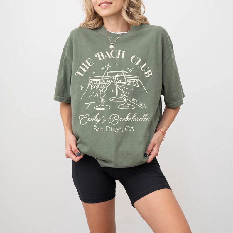 May include: Green t-shirt with a white graphic design that says "The Bach Club" with a martini glass design and the text "Emily's Bachelorette San Diego, CA".