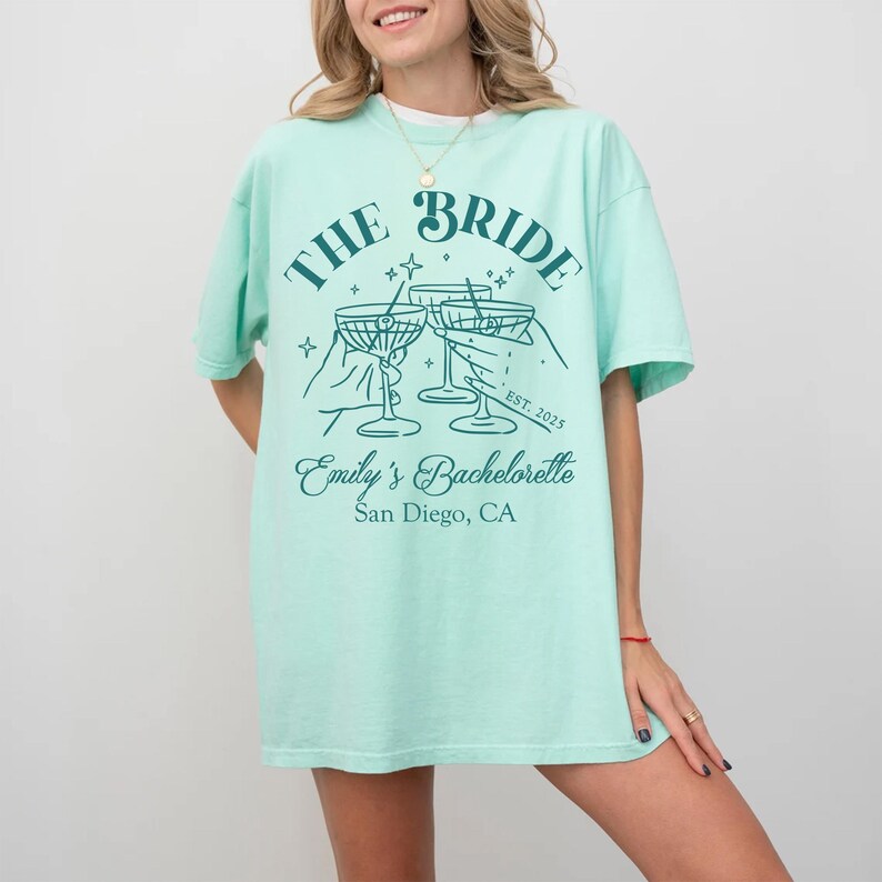 May include: A light green t-shirt with the text "The Bride" in a decorative font. The shirt also features a graphic of three martini glasses with the text "Emily's Bachelorette San Diego, CA" below the glasses.