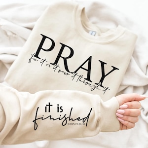 Op de afbeelding: Een beige sweatshirt met de tekst "PRAY for it, on it, over it, through it" op de voorkant en "it is finished JOHN 19:28-30" op de mouw.