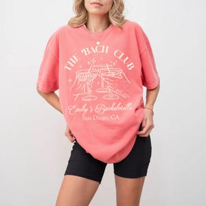May include: A coral pink t-shirt with a white graphic design that says "The Bach Club" with a martini glass graphic and the text "Emily's Bachelorette San Diego, CA"