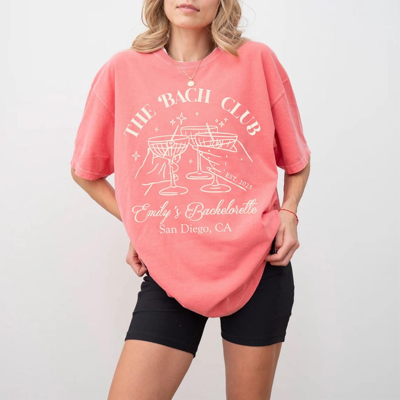 May include: A coral pink t-shirt with a white graphic design that says "The Bach Club" with a martini glass graphic and the text "Emily's Bachelorette San Diego, CA"