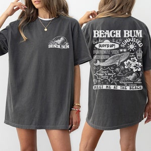 May include: Dark grey t-shirt with the text "BEACH BUM" on the front and back. The back of the shirt features a graphic with the words "SURFS UP! SUNKISSED" and other beach-themed illustrations, including a whale, sunglasses, and palm trees.
