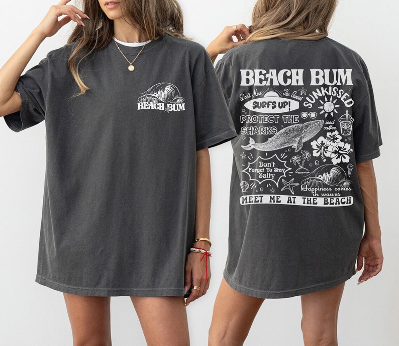 May include: Dark grey t-shirt with the text "BEACH BUM" on the front and back. The back of the shirt features a graphic with the words "SURFS UP! SUNKISSED" and other beach-themed illustrations, including a whale, sunglasses, and palm trees.