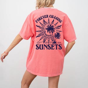 Puede incluir: Una camiseta de color rosa coral con un gr&aacute;fico azul marino en la espalda. El gr&aacute;fico presenta un sol con palmeras y el texto "FOREVER CHASING SUNSETS".