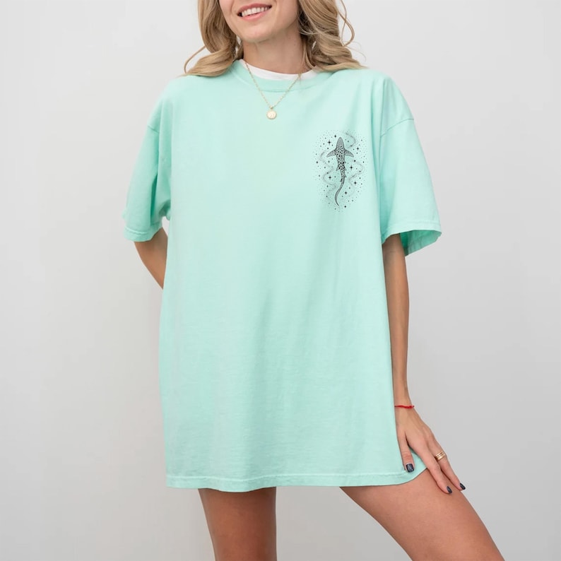 Puede incluir: Camiseta verde claro con una peque&ntilde;a ilustraci&oacute;n en blanco y negro de un pez en la parte superior derecha del pecho. La camiseta es holgada y tiene mangas cortas.