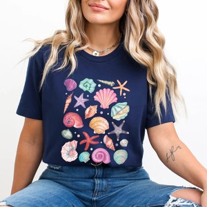 K&ouml;nnte beinhalten: Ein marineblaues T-Shirt mit einem farbenfrohen Design aus Muscheln und Seesternen. Das Design ist mittig auf dem Shirt platziert und zeigt eine Vielzahl verschiedener Muscheln in den Farben Rosa, Lila, Blau und Gr&uuml;n.