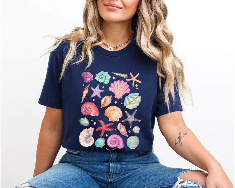 K&ouml;nnte beinhalten: Ein marineblaues T-Shirt mit einem farbenfrohen Design aus Muscheln und Seesternen. Das Design ist mittig auf dem Shirt platziert und zeigt eine Vielzahl verschiedener Muscheln in den Farben Rosa, Lila, Blau und Gr&uuml;n.