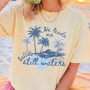 May include: A light yellow t-shirt with a palm tree graphic and the text "He leads me beside still waters". The shirt is worn on a beach with a sunset in the background. A person is holding a green drink.