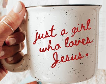 Op de afbeelding: Witte gespikkelde keramische mok met de tekst "just a girl who loves Jesus" in rood script. De mok wordt in een hand gehouden, met een boek en andere items op de achtergrond.