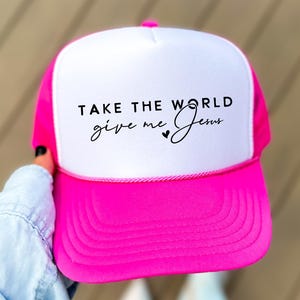 May include: A bright pink trucker hat with a white front panel. The hat features the text "TAKE THE WORLD give me Jesus" in black script. The hat has a curved brim and a mesh back.