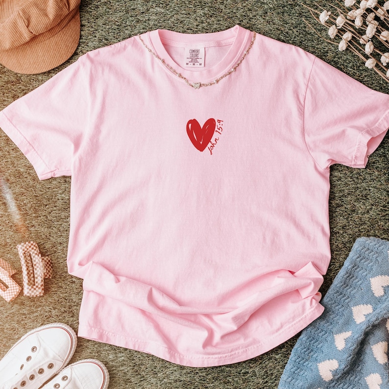 Op de afbeelding: Roze T-shirt met een rood hart en de tekst "Johannes 15:9" op de voorkant.