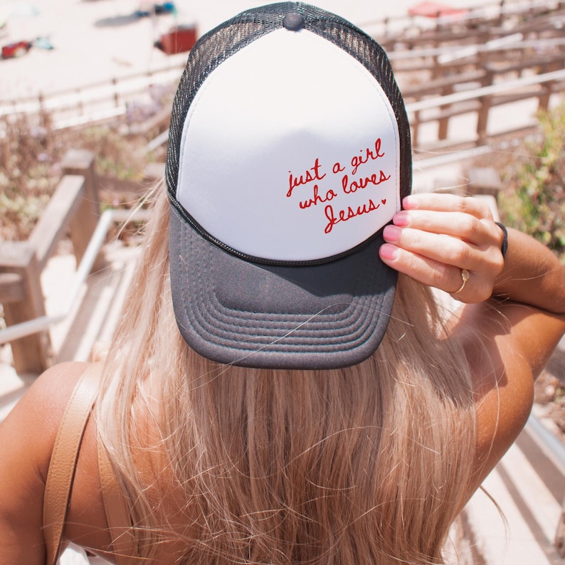 Op de afbeelding: Een witte trucker-pet met een zwart mesh-achterpaneel. De pet heeft rode geborduurde tekst die "just a girl who loves Jesus." luidt.