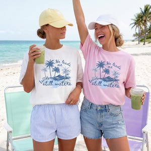 K&ouml;nnte beinhalten: Zwei Frauen tragen T-Shirts mit einem Strandmotiv und dem Text "He leads me beside still waters". Ein Shirt ist cremefarben, das andere rosa. Eine Frau tr&auml;gt eine gelbe Kappe, die andere eine wei&szlig;e Kappe. Beide halten Getr&auml;nke.