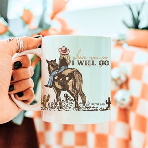 May include: White ceramic mug with a brown and white horse and a woman in a pink hat riding it. The mug has the text "where you go I WILL GO" and "RUTH 1:16" printed on it.