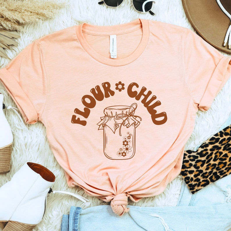 May include: A light pink t-shirt with a brown graphic of a jar with flowers and the text "Flour Child" in a retro font.