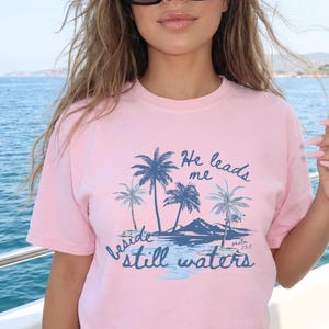 K&ouml;nnte beinhalten: Hellrosa T-Shirt mit blauem Aufdruck von Palmen, Bergen und dem Text "He leads me beside still waters psalm 23:2". Das T-Shirt hat einen Rundhalsausschnitt und kurze &Auml;rmel.