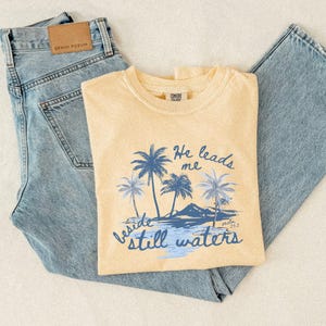 K&ouml;nnte beinhalten: Hellgelbes T-Shirt mit blauem Palmen-Motiv und dem Text "He leads me beside still waters". Das Shirt wird mit hellblauen Jeans kombiniert. Das Shirt hat ein Comfort Colors-Etikett.