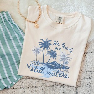 K&ouml;nnte beinhalten: Cremefarbenes T-Shirt mit blauem Palmen-Motiv und dem Text "He leads me beside still waters." Eine goldene Halskette und gr&uuml;ne gestreifte Shorts sind ebenfalls zu sehen. Das Shirt ist von der Marke Comfort Colors.