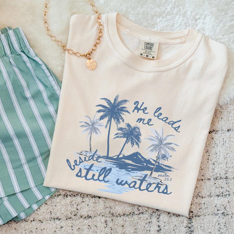 K&ouml;nnte beinhalten: Cremefarbenes T-Shirt mit blauem Palmen-Motiv und dem Text "He leads me beside still waters." Eine goldene Halskette und gr&uuml;ne gestreifte Shorts sind ebenfalls zu sehen. Das Shirt ist von der Marke Comfort Colors.