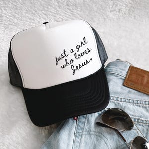 May include: A white and black trucker hat with the text "just a girl who loves Jesus" in black script. The hat has a black brim and mesh back. Sunglasses and denim jeans are in the background.
