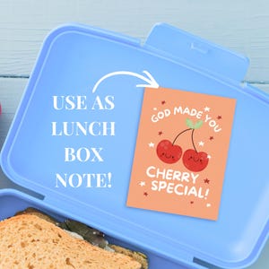 Peut inclure: Une bo&icirc;te &agrave; lunch bleu clair avec un sandwich &agrave; l'int&eacute;rieur. Une petite carte orange avec le texte "GOD MADE YOU CHERRY SPECIAL!" et deux cerises souriantes est &agrave; l'int&eacute;rieur. Le texte "USE AS LUNCH BOX NOTE!" est &eacute;galement visible.
