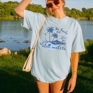 K&ouml;nnte beinhalten: Hellblaues T-Shirt mit einer Grafik von Palmen, Bergen und dem Text "He leads me beside still waters". Das Shirt hat eine lockere Passform und kurze &Auml;rmel. Im Hintergrund sind ein See und gr&uuml;ne B&auml;ume zu sehen.