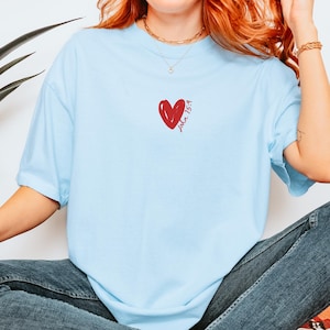 Op de afbeelding: Een lichtblauw t-shirt met een rood hart en de tekst "Johannes 15:9" op de voorkant.