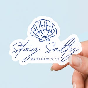 Puede incluir: Una pegatina blanca con un diseño de concha azul y la frase "Stay Salty" en cursiva. El texto "Matthew 5:13" está debajo. La pegatina se sostiene sobre un fondo azul claro.