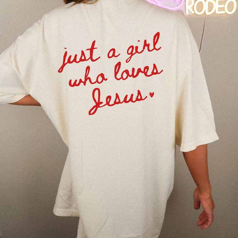 Op de afbeelding: Een wit oversized T-shirt met de tekst "just a girl who loves Jesus" in rode cursieve letters.