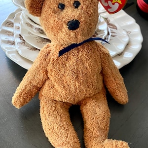 May include: A plush brown teddy bear with black eyes and a black nose. The bear has a blue ribbon tied around its neck and a tag attached to its ear. The bear is sitting in front of a stack of white plates.