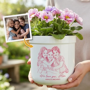 May include: A white ceramic flower pot with a pink line drawing of a family hugging. The pot contains pink and purple flowers. The text "We love you, Mom" and names are printed on the pot.