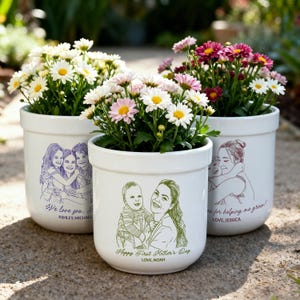 May include: Three white ceramic flower pots filled with colorful flowers. Each pot features a line drawing of a family or individual with text. The flowers include white, pink, and maroon blooms. The pots are sitting on a stone surface.
